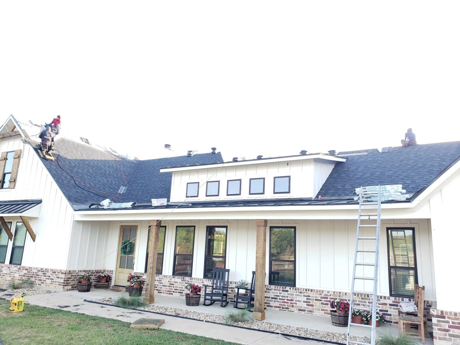 Two roofers actively installing asphalt shingles on a large white farmhouse-style home under a bright sky